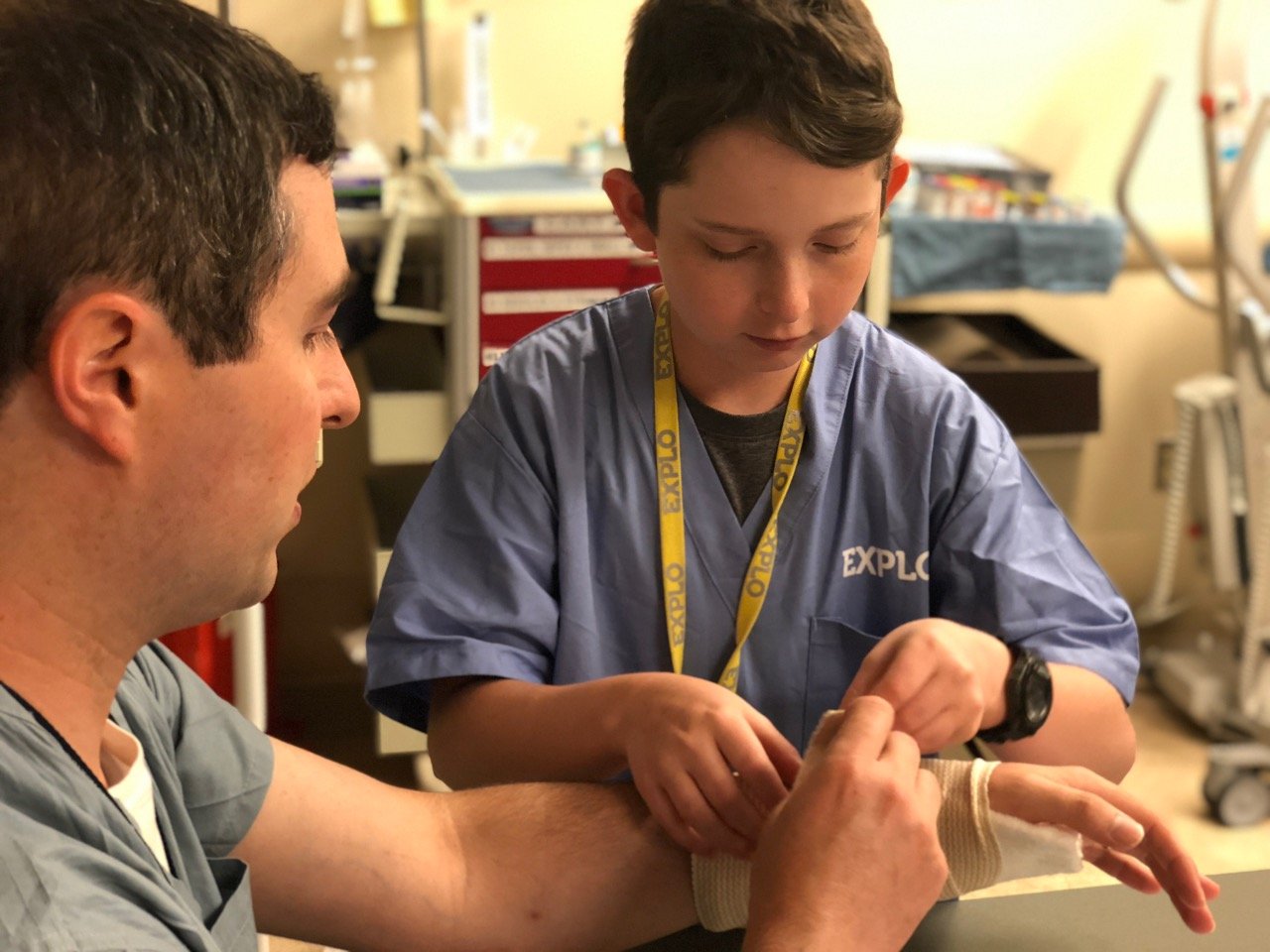 Dr. Eyre sits as a young male EXPLO ER student wraps his wrist in an ACE bandage